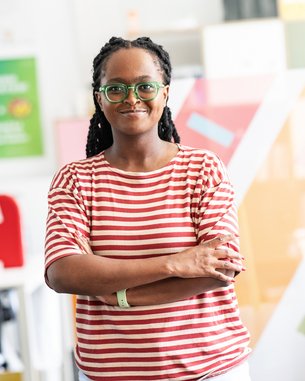 Eine Frau mit langen, geflochtenen Haaren trägt ein gestreiftes T-Shirt und steht mit verschränkten Armen in einem modernen Büro. Sie trägt eine Brille und lächelt leicht, während bunte Wände im Hintergrund sichtbar sind.