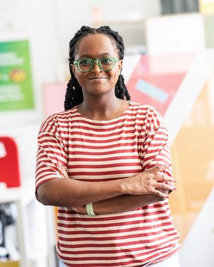 Eine Frau mit langen, geflochtenen Haaren trägt ein gestreiftes T-Shirt und steht mit verschränkten Armen in einem modernen Büro. Sie trägt eine Brille und lächelt leicht, während bunte Wände im Hintergrund sichtbar sind.