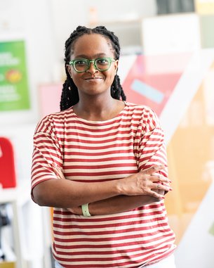Eine Frau mit langen, geflochtenen Haaren trägt ein gestreiftes T-Shirt und steht mit verschränkten Armen in einem modernen Büro. Sie trägt eine Brille und lächelt leicht, während bunte Wände im Hintergrund sichtbar sind.