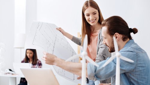 Zwei Personen besprechen Pläne, während eine dritte Person am Computer arbeitet. Auf dem Tisch stehen kleine Windturbinenmodelle.