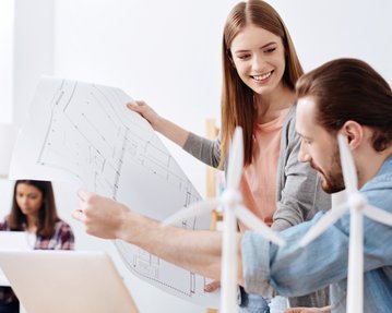 Zwei Personen besprechen Pläne, während eine dritte Person am Computer arbeitet. Auf dem Tisch stehen kleine Windturbinenmodelle.