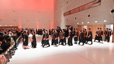 Graduates in academic robes walk through a festive event hall during a graduation ceremony.
