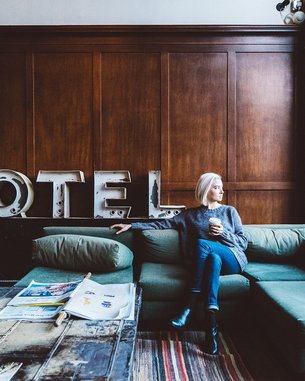 Eine Frau mit blonden Haaren sitzt auf einem grünen Sofa in einem Hotel-Lobby. Sie hält eine Tasse in der Hand und blickt nachdenklich aus dem Fenster. Im Hintergrund steht das Wort "HOTEL" in großen Buchstaben.