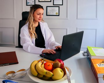 Eine Frau in einem weißen Kittel sitzt an einem Schreibtisch und arbeitet an einem Laptop. Auf dem Tisch befindet sich eine Schüssel mit Obst und ein Maßband. Im Hintergrund hängen Zertifikate an der Wand.
