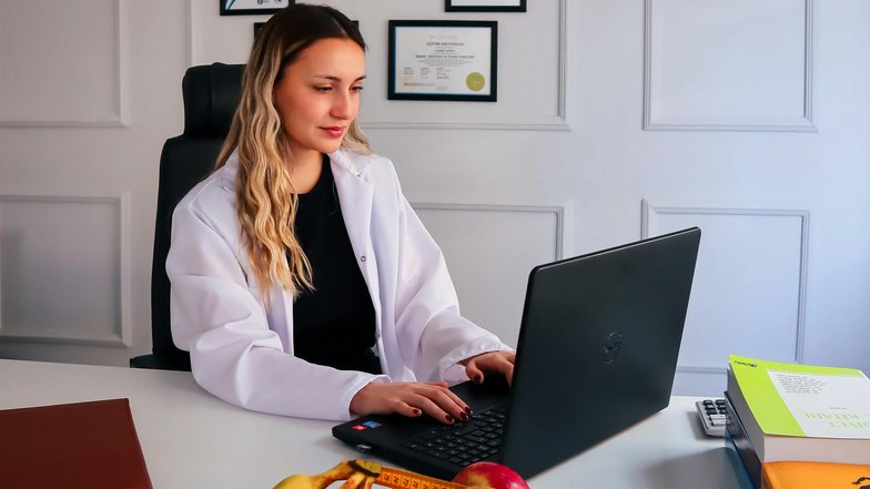 Eine Frau in einem weißen Kittel sitzt an einem Schreibtisch und arbeitet an einem Laptop. Auf dem Tisch befindet sich eine Schüssel mit Obst und ein Maßband. Im Hintergrund hängen Zertifikate an der Wand.