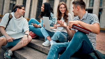 Four young adults are sitting on steps, chatting and using laptops and notebooks. They appear relaxed and friendly.