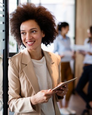 Eine Frau mit lockigem Haar steht an einer Glaswand und hält ein Tablet. Sie trägt einen blassen Blazer und lächelt. Im Hintergrund sind zwei weitere Frauen zu sehen, die miteinander sprechen.