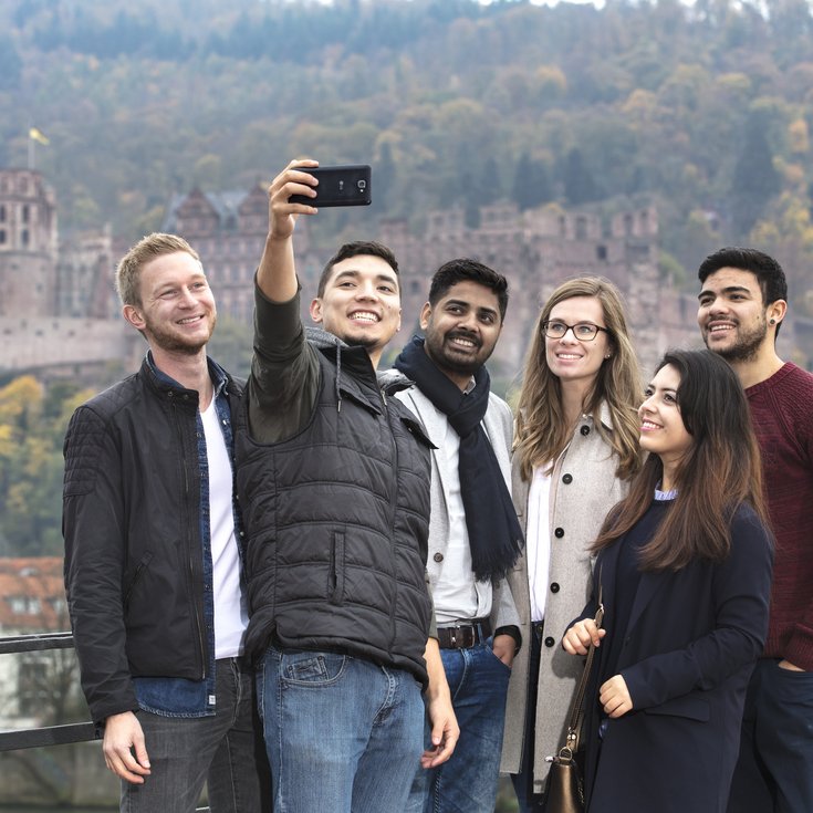 Six people are taking a selfie in front of a historic castle and an autumn landscape.
