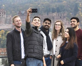 Six people are taking a selfie in front of a historic castle and an autumn landscape.
