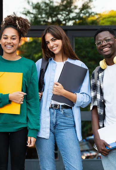 Four students are smiling in front of a building, carrying backpacks and holding books and documents in their hands.