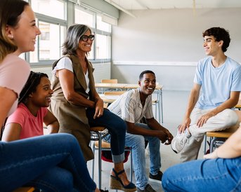 Six people are sitting in a classroom and having a conversation. One woman is standing while the others are sitting in chairs and smiling.