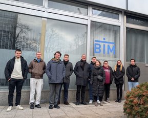 Eine Gruppe von acht Personen steht vor einem Gebäude mit dem Logo "BIM". Sie tragen verschiedene Winterkleidung und posieren freundlich.