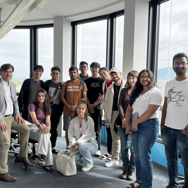 Group of 16 people in a modern office with large windows, some sitting, others standing, all smiling and posing for a photo.