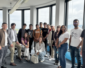 Gruppe von 16 Personen in einem modernen Büro mit großen Fenstern, einige sitzen, andere stehen, alle lächeln und posieren für ein Foto.