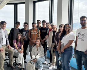 Group of 16 people in a modern office with large windows, some sitting, others standing, all smiling and posing for a photo.