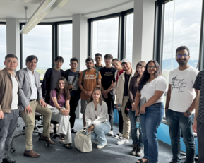 Group of 16 people in a modern office with large windows, some sitting, others standing, all smiling and posing for a photo.