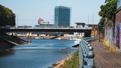 Uferpromenade entlang eines Flusses mit einer Brücke im Hintergrund und modernen Gebäuden in der Ferne.