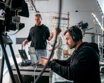 A man with headphones is working on a laptop while another man gestures in the background. Camera equipment and lighting are visible.