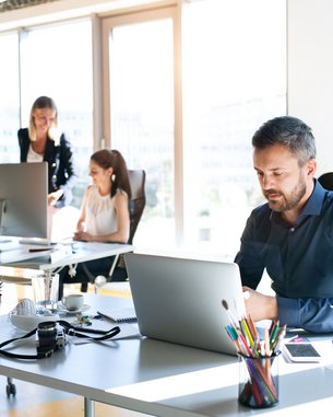 Drei Personen arbeiten in einem modernen Büro. Ein Mann sitzt an einem Schreibtisch und nutzt einen Laptop, während zwei Frauen an einem anderen Tisch beschäftigt sind. Pflanzen und Büromaterialien sind sichtbar.