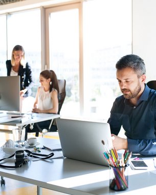 Drei Personen arbeiten in einem modernen Büro. Ein Mann sitzt an einem Schreibtisch und nutzt einen Laptop, während zwei Frauen an einem anderen Tisch beschäftigt sind. Pflanzen und Büromaterialien sind sichtbar.