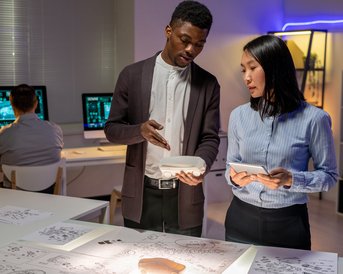 Two people are discussing plans while holding tablets in a modern office, while a third person is working at a computer.