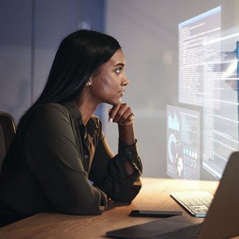 A woman sits at a desk, intently looking at several computer screens filled with programming code and data visualizations.