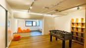 Ein heller Raum mit Holzboden, einem Tischfußballspiel in der Mitte und orangefarbenen Sitzsäcken in einer Ecke.