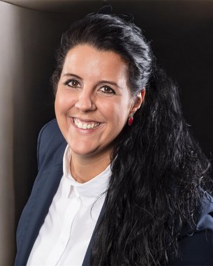 Eine Frau mit langen, schwarzen Haaren und einem blauen Blazer lächelt in die Kamera, während sie in einem hellen Raum steht.