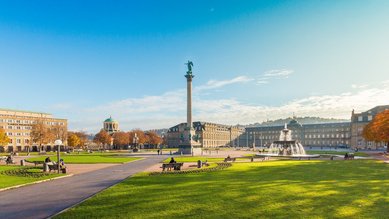 A spacious park with a monument in the center, surrounded by historic buildings and a fountain. The sun is shining, and the sky is clear. Benches invite you to sit and relax.