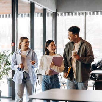 Three young adults are chatting in a café while holding notebooks and a coffee cup.