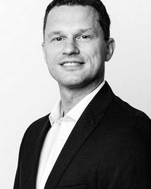 Ein Mann mit kurzem Haar trägt einen schwarzen Blazer und ein weißes Hemd. Er lächelt und steht vor einem neutralen Hintergrund.