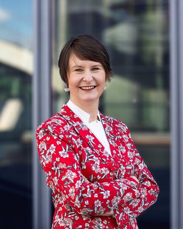 Frau mit kurzem Haar trägt einen roten Blazer mit Blumenmuster und steht lächelnd vor einer modernen Glasfassade.