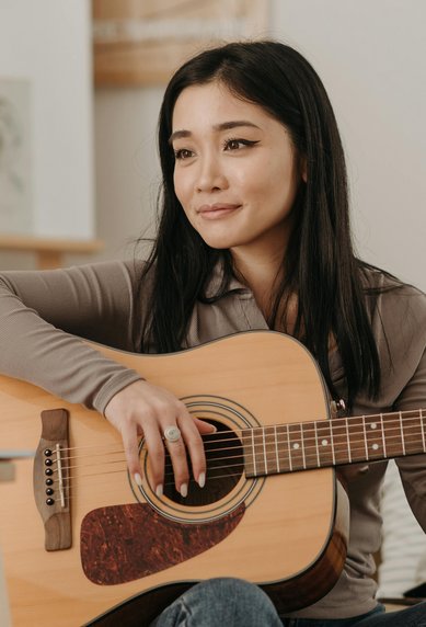Eine Frau mit langen, schwarzen Haaren spielt Gitarre auf einem Sofa in einem hellen Raum. Sie trägt ein graues Oberteil und wirkt konzentriert.