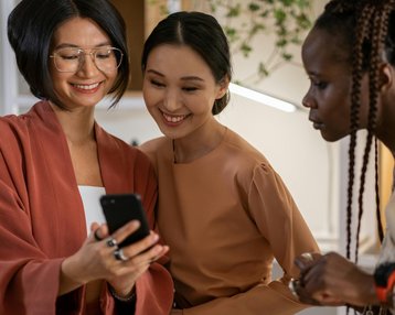 Drei Frauen stehen zusammen und schauen auf ein Smartphone. Eine Frau mit Brille lächelt, während die anderen beiden interessiert auf den Bildschirm blicken. Sie tragen modische, farblich abgestimmte Kleidung.