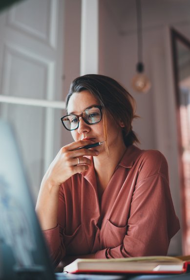 Eine Frau mit Brille sitzt an einem Tisch, schaut nachdenklich auf ihren Laptop und hält einen Stift in der Hand.