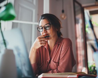 A woman with glasses sits at a table, thoughtfully looking at her laptop while holding a pen in her hand.