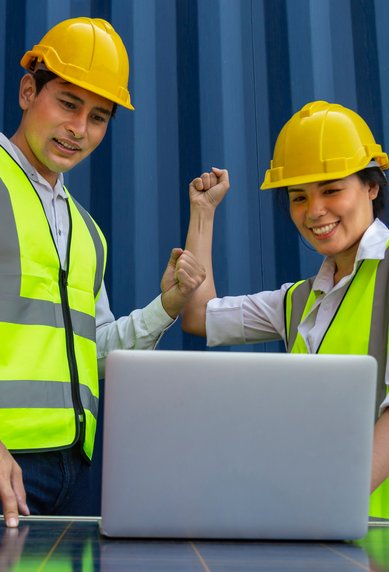 Two engineers in safety vests and helmets are looking at a laptop while they celebrate wind turbines.