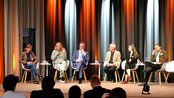 Eine Diskussionsrunde mit sechs Personen auf einer Bühne, die in einem modernen Raum sitzen. Die Teilnehmer sind in formeller Kleidung und diskutieren, während einige Wasserflaschen auf den Tischen stehen.