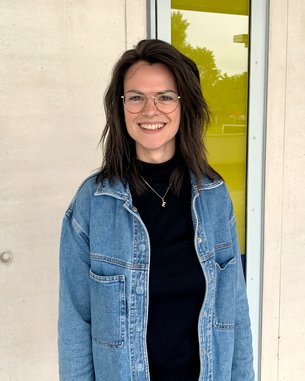 Eine junge Frau mit Brille und lockigem Haar trägt eine Jeansjacke und steht vor einer farbigen Wand. Sie lächelt freundlich.