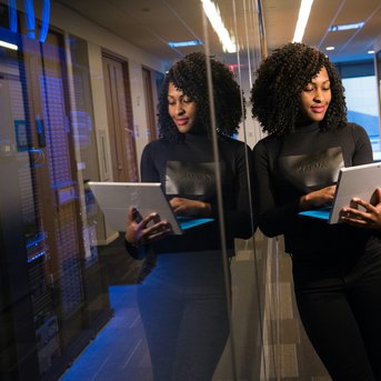 A woman with curly hair stands in a modern office, looking at a tablet. She is in front of a mirror, reflecting in a room with servers in the background.