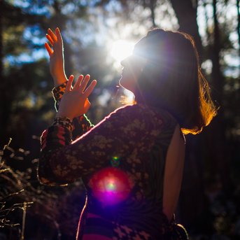 A person with long hair stands in the forest, raising their hands to the sun that shines through the trees. They are wearing a colorful dress.