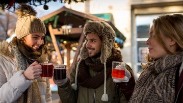 Three people toast with mulled wine and other drinks while enjoying themselves in a festively decorated outdoor area.