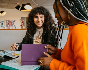 Zwei junge Frauen sitzen an einem Tisch, umgeben von Schulmaterialien. Eine Frau mit lockigem Haar lächelt, während die andere mit geflochtenen Haaren ein lila Heft hält. Im Hintergrund sind mehrere Trophäen sichtbar.