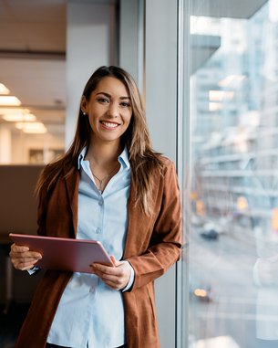 Eine lächelnde Frau steht an einem Fenster in einem modernen Bürogebäude und hält ein Tablet in der Hand. Sie trägt eine braune Jacke und ein hellblaues Hemd. Im Hintergrund sind Stadtansichten und Büroeinrichtungen sichtbar.
