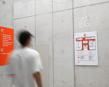 Signpost and floor plan on the ground floor of a building, with information about design studios and labs. A person walks by.