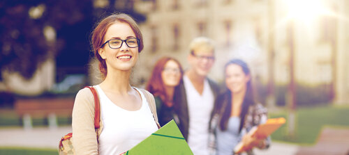 A smiling student with glasses is holding a green notebook, while three other students stand in the background.