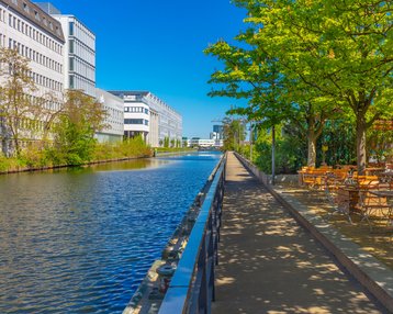 A paved path along a canal, lined with trees and outdoor seating, with modern buildings in the background.