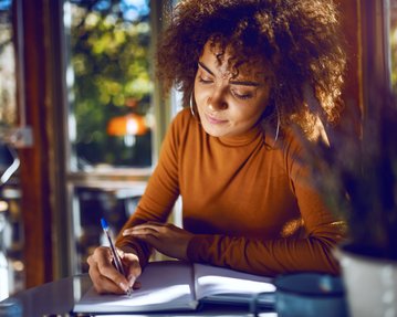 A woman with curly hair sits at a table, writing in a notebook while working in a bright café.