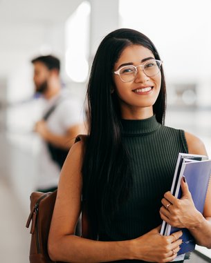 Eine junge Frau mit langen, schwarzen Haaren und Brille hält Bücher in der Hand und lächelt, während sie in einem modernen Lernumfeld steht.