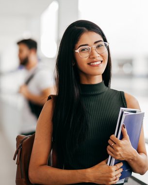 Eine junge Frau mit langen, schwarzen Haaren und Brille hält Bücher in der Hand und lächelt, während sie in einem modernen Lernumfeld steht.